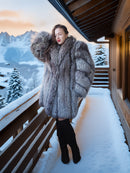 Person wearing a fur coat standing on a snowy balcony with mountains in the background