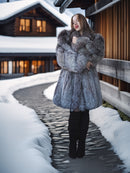 Woman in a fur coat standing on a snowy path in front of a wooden building.