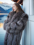 Woman wearing a fur coat standing by a window with a mountain view.