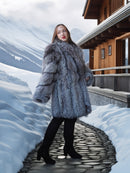 Person wearing a fur coat standing on a snowy path near a wooden cabin.