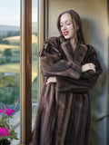 Woman wearing a brown fur coat standing by a window with a scenic view.