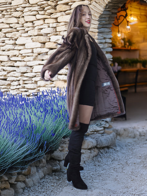 Woman in a fur coat standing next to a stone wall with lavender plants.