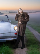 Woman in a fur coat standing next to a vintage car on a foggy road.