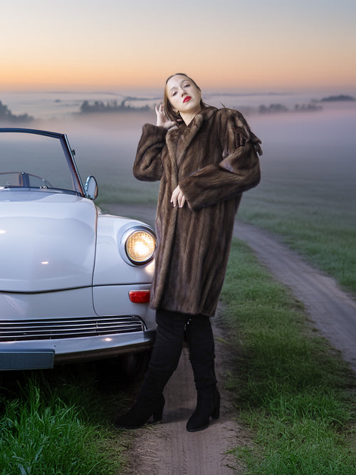 Woman in a fur coat standing next to a vintage car on a foggy road.