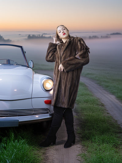 Woman in a fur coat standing next to a vintage car on a foggy road.