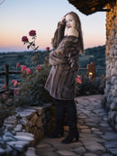 Woman in a fur coat standing on a stone path with flowers and a sunset in the background