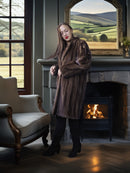 Woman in a fur coat standing in a room with a fireplace and a large mirror showing a scenic view.