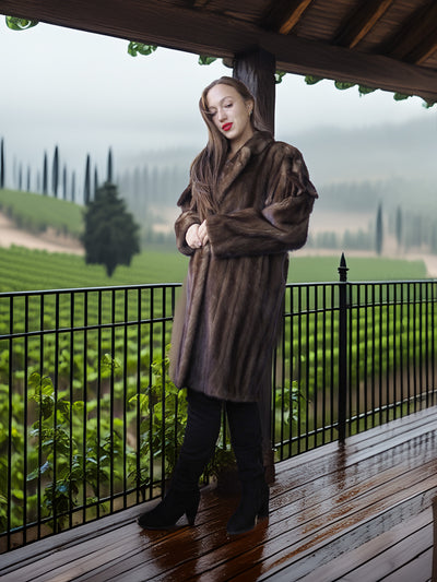Woman in a brown fur coat standing on a wooden deck with vineyard view