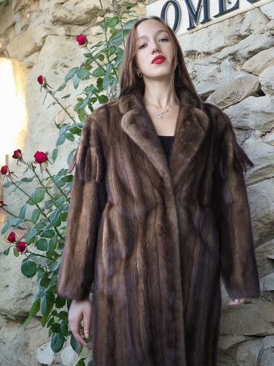 Woman wearing a brown fur coat standing against a stone wall with red flowers.