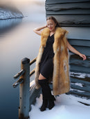 Woman in a fur vest standing by a snowy landscape with water and a building.