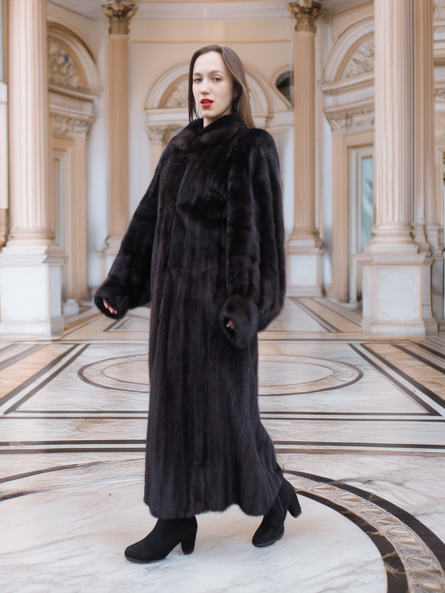 Model walks confidently in full-length black mink coat, styled for upscale winter fashion in a neoclassical setting.