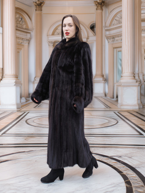 Model walks confidently in full-length black mink coat, styled for upscale winter fashion in a neoclassical setting.