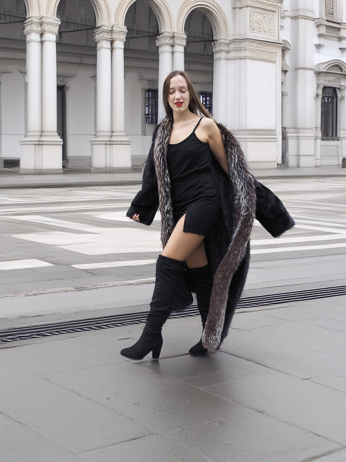 Model in black mink coat with indigo fox trim, walking confidently in a city square, styled with black boots.
