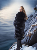 Person wearing a fur coat standing on a snowy dock by a body of water.