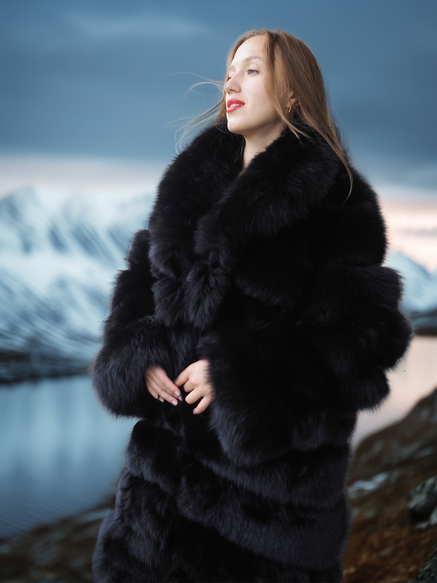 Woman wearing a black fur coat standing in a scenic outdoor setting with mountains and water.
