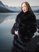 Woman wearing a black fur coat standing in front of a lake with mountains in the background