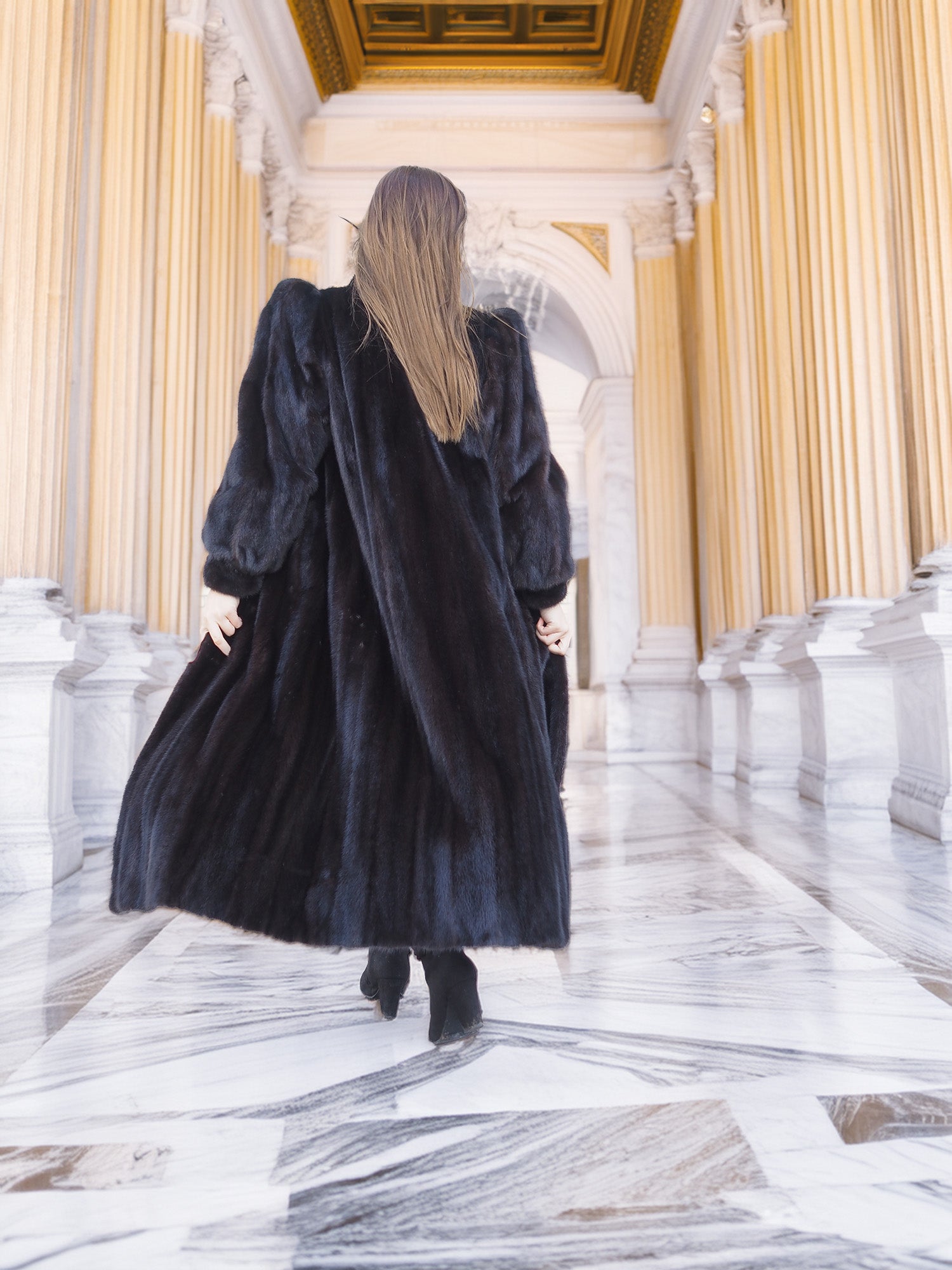 Back view of black mink fur coat with dramatic volume and soft texture, modeled in a marble corridor. Luxury outerwear, size M.