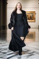 Woman in a black coat and dress standing in an elegant interior setting.