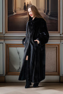 Woman wearing a long black coat standing in an elegant interior setting with framed pictures on the wall.