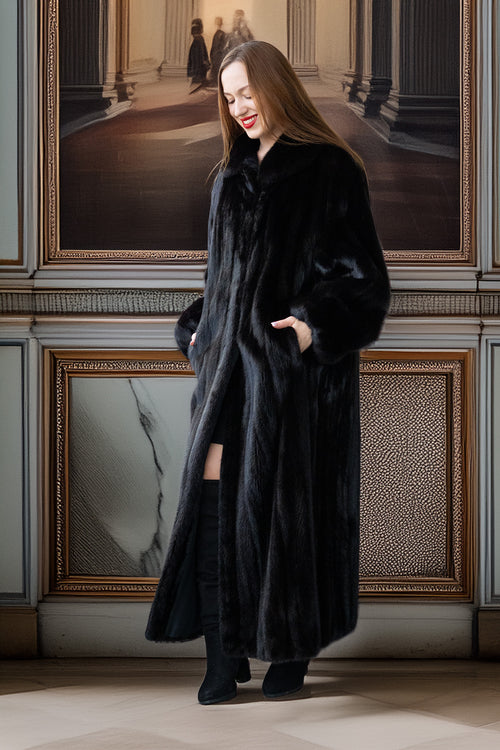Woman wearing a long black coat standing in an elegant interior setting with framed pictures on the wall.