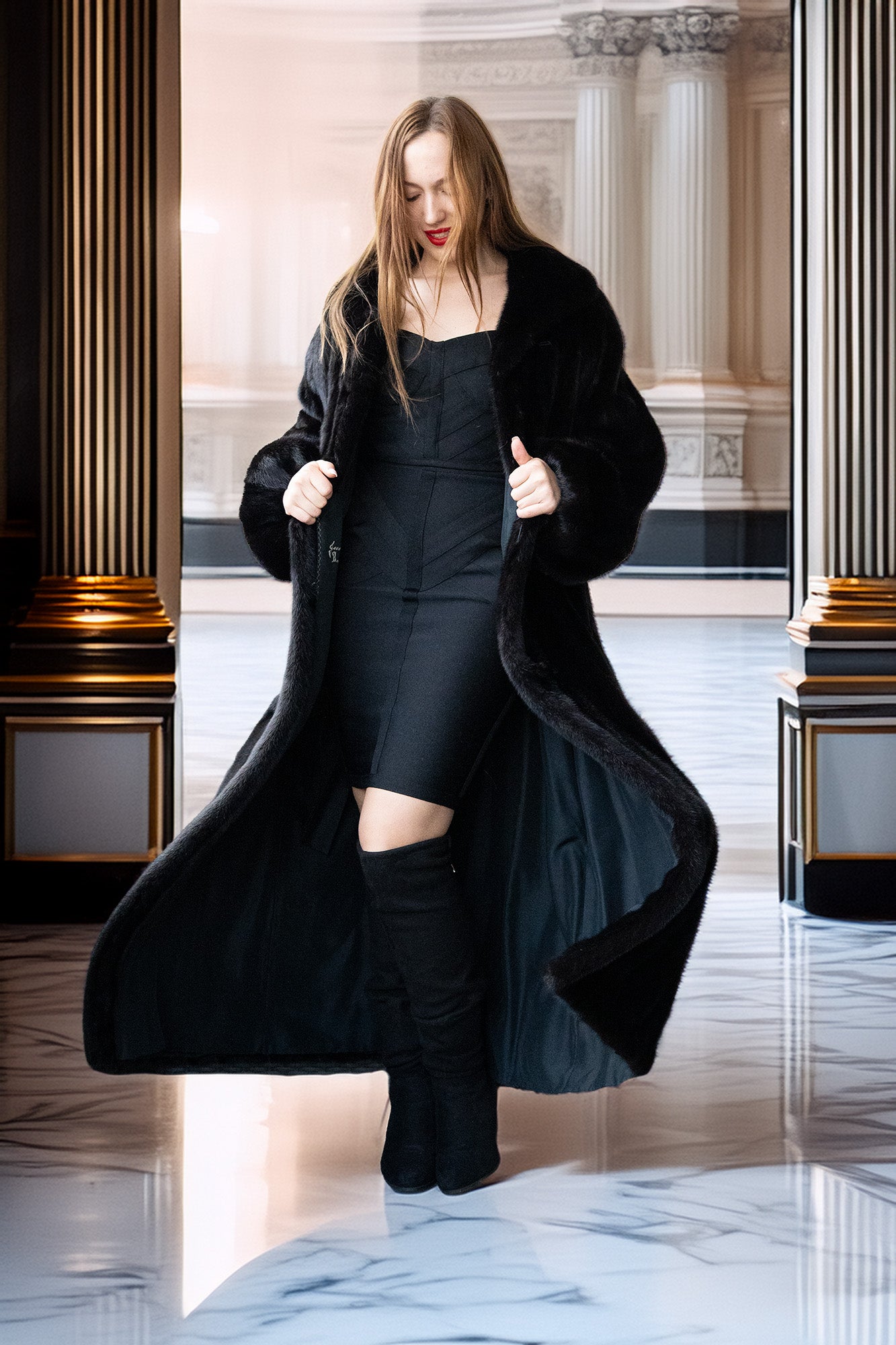 Woman in a long black coat standing in a grand hallway.