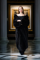 Woman in a black dress standing in a room with classical architecture and a painting in the background.