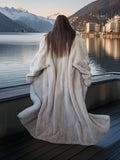 Person in a fur coat standing by a lake with mountains in the background