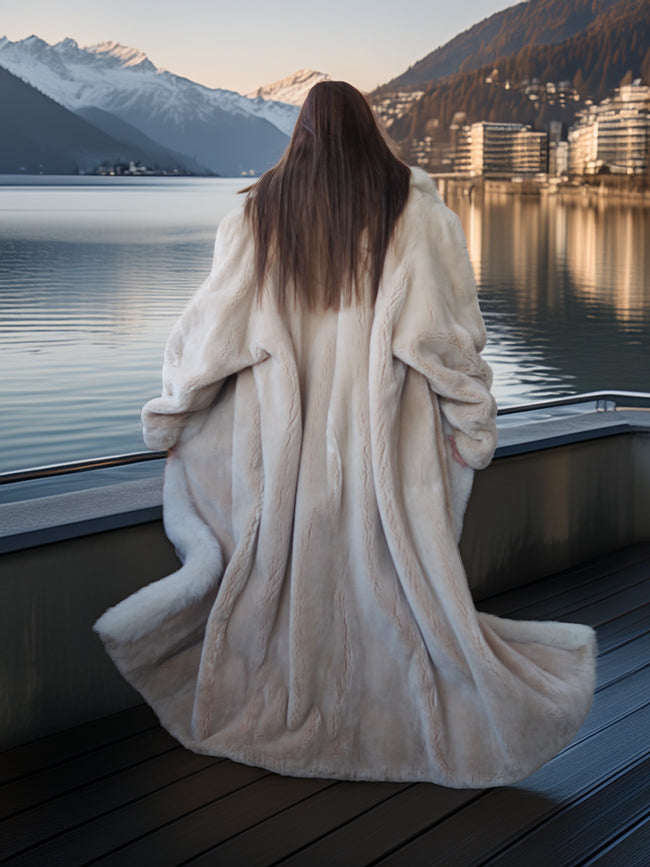 Person in a fur coat standing by a lake with mountains in the background