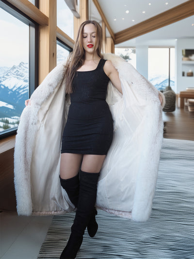 Woman wearing a long white fur coat in a modern interior setting with large windows.