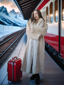 Woman in a fur coat with a red suitcase standing on a train platform with mountains in the background