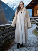 Woman in a fur coat standing in a mountainous area with a wooden cabin.