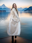 Woman in a fur coat standing by a lake with mountains in the background