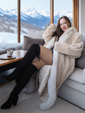 Woman in a fur coat sitting on a couch with a mountain view outside the window.