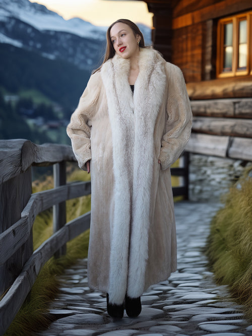 Woman in a long fur coat standing in front of a wooden cabin with a mountainous background