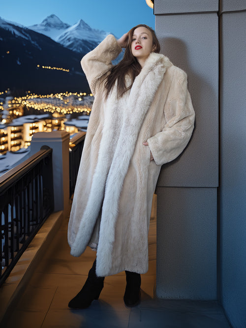 Woman in a long white fur coat standing on a balcony with a cityscape and mountains in the background.
