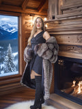 Woman in a fur coat standing in a cozy room with a fireplace and snowy landscape outside.