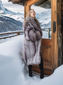 Woman in a fur coat standing in front of a wooden door with a snowy mountain landscape.