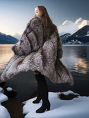 Person wearing a fur coat standing by a snowy landscape with mountains and water.