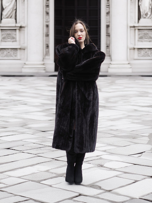 Model poses in a full-length black mink fur coat with arms crossed, showcasing its plush texture and elegant fit.