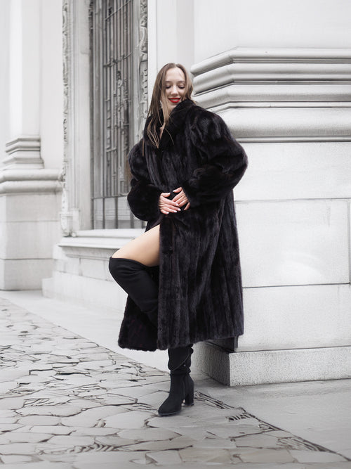 Model leans against a wall in a black mink fur coat, styled with thigh-high boots for a modern luxury look.
