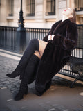 Woman in a long black fur coat and knee-high boots sitting on a bench in an urban setting.