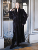 Woman in a long black coat standing in an elegant interior setting.