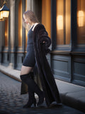 Woman in a long coat and knee-high boots standing on a street with a building in the background.