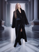 Woman in a long black coat standing in a grand hallway with columns.