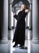 Woman in a long black coat standing in a classical architectural setting with columns.