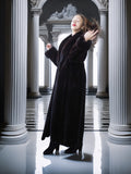 Woman in a long black coat standing in a classical architectural setting with columns.