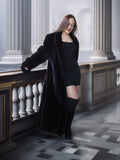 Woman in a long black coat and black dress standing in an elegant interior setting.