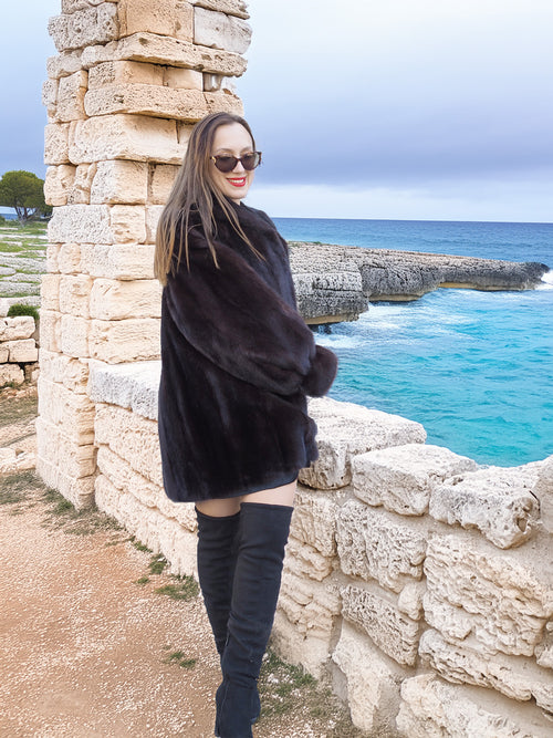 Black Mink Swing Coat - Riviera Noir worn near a stone wall overlooking the sea. Elegant fur outerwear for cold-weather sophistication.