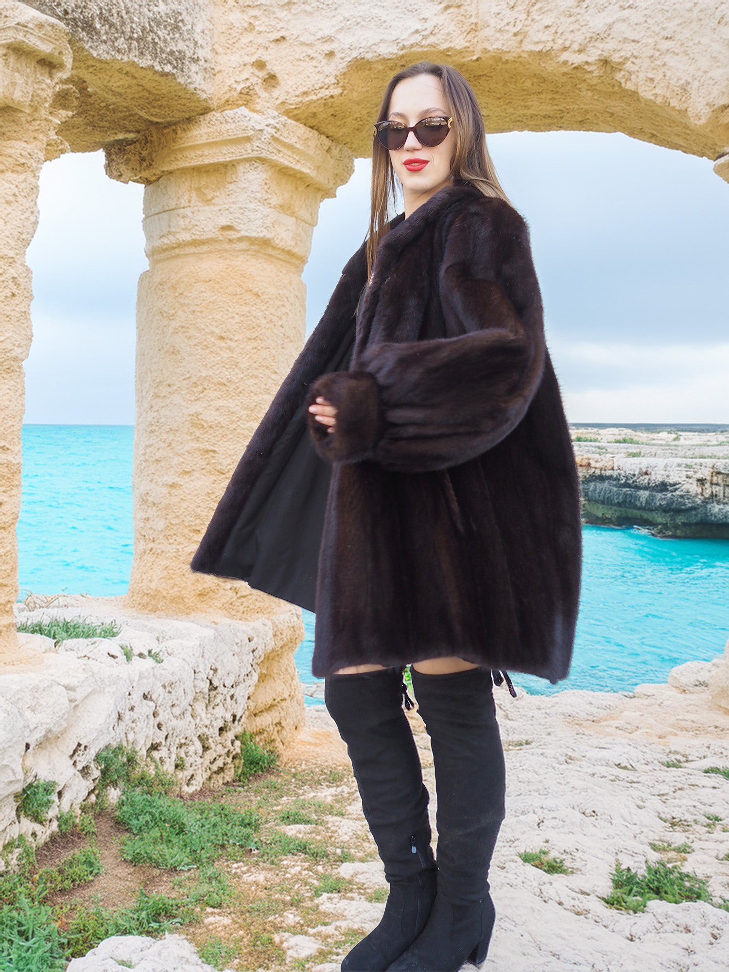 Riviera Noir Black Mink Swing Coat styled near ancient stone arches. Premium mink coat for timeless winter fashion.