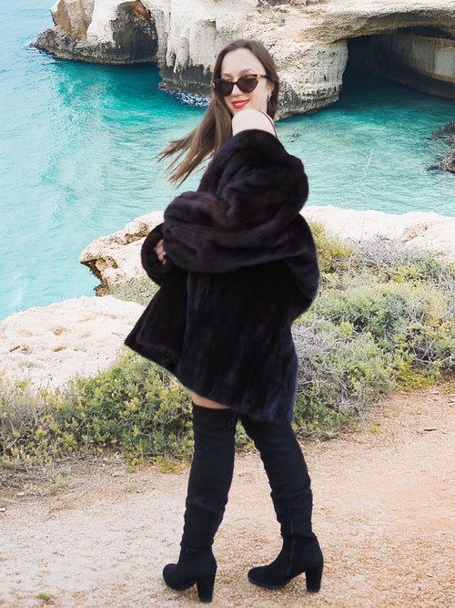 Black Mink Swing Coat - Riviera Noir in motion with a scenic ocean backdrop. Elegant and warm fur coat for luxury winter wear.